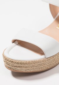 Valkoinen nahkainen korkokenkä leveällä hihnalla, juuttikutoisella pohjalla ja sileällä pinnalla. Sisältää pyöristetyn varpaan ja hillityn tikkausyksityiskohdan.