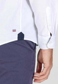 Chemise à manches longues blanche avec des poignets à boutons, ornée d'une accentuation bleu marine à l'ourlet et d'un petit patch logo rouge et bleu marine près du bas.