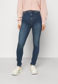 Jeans skinny de cintura alta azul em tecido de denim com uma textura suave e costura laranja, combinados com ténis brancos chunky.
