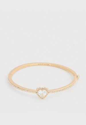 Arany karperec szív alakú cirkónia központtal és apró tiszta kristályokkal a szíjon, sima textúrájú felülettel.