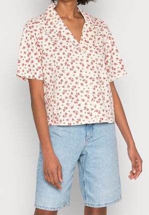 Personne portant une chemise crème à manches courtes avec un petit motif floral rouge et un short en jean bleu clair jusqu'aux genoux.