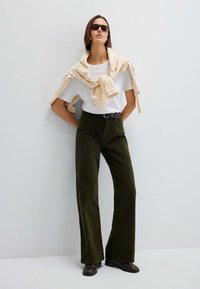 Pantaloni di velluto a coste verde scuro, maglietta bianca, maglione beige drappeggiato sulle spalle, e occhiali da sole neri. Texture morbida e design a gamba larga.