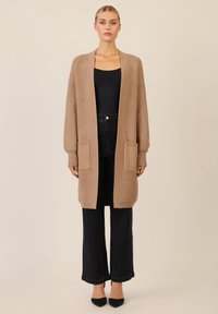 Cardigan lungo con apertura frontale in marrone chiaro, texture a coste, dotato di due tasche frontali, abbinato a jeans scuri a vita alta e tacchi neri.