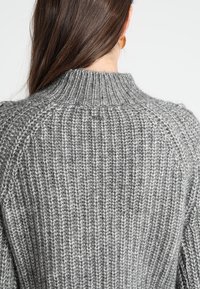 De longs cheveux bruns tombent sur un gros pull gris en maille avec un col montant côtelé, vu de dos.