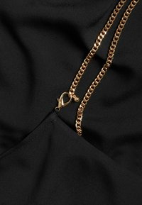 Svart tyg med en slät yta, med detaljer i guld kedja och spänne. Designen inkluderar en triangulär halsringning och dragkedja.