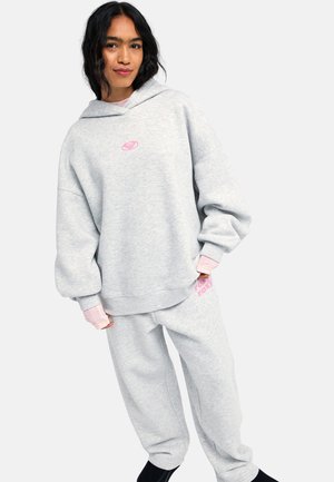 Frau trägt einen hellgrauen oversized Hoodie und passende Jogginghose mit pinkfarbenen Logodetails, stehend vor schlichtem Hintergrund.