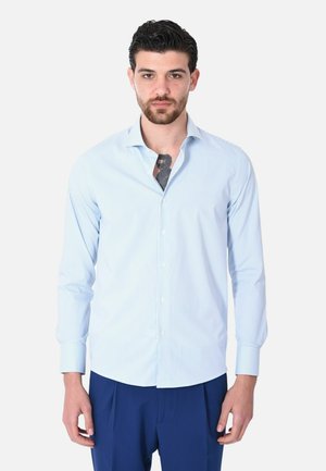 Uomo con capelli scuri e barba che indossa una camicia azzurro chiaro a righe parzialmente sbottonata che mostra un tatuaggio sul petto e pantaloni blu scuro.