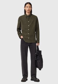 Mand iført en olivengrøn skjorte med knapper og sorte jeans, holder en sort quilted taske i remmen, mens han står mod en ensfarvet baggrund.