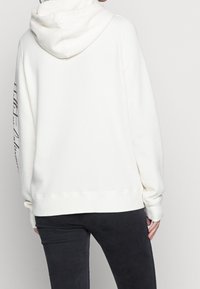 Weißer Kapuzenpullover aus weichem, strukturiertem Stoff; mit gerippten Bündchen und Saum; Druckdetails auf dem linken Ärmel.