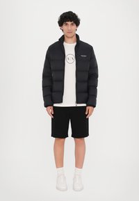 Sort pufferjakke med lynlås foran, der har høj krave og syede paneler. Bæres over en hvid t-shirt og sorte shorts, med hvide sneakers.