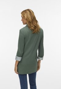 Zielona marynarka o dopasowanym kroju, z podwiniętymi paskami w paski. Gładki materiał i usystematyzowany design, pokazana z tyłu.