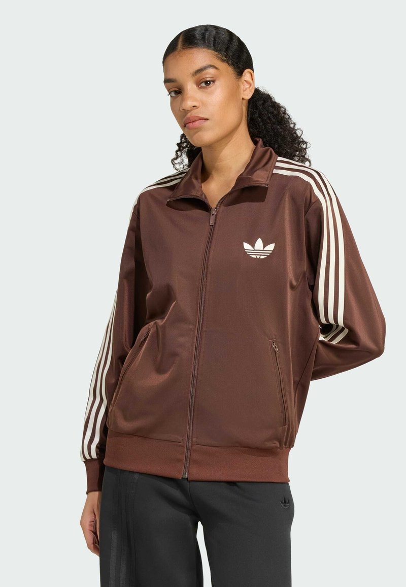 Veste de sport zippée marron en matériau lisse et brillant, avec des détails à trois bandes blanches sur les manches et un petit logo sur la poitrine.