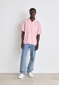 Kortermet rosa stripete skjorte med lomme, kombinert med lyseblå rette jeans og hvite sneakers, stående på en nøytral bakgrunn.