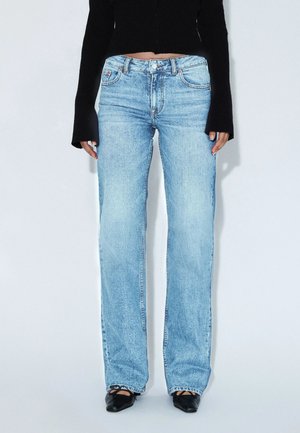 Person trägt hellblaue Jeans mit geradem Bein, schwarzen kurzen Pullover und schwarze spitze Schuhe mit Knöchelriemen, steht vor weißem Hintergrund.