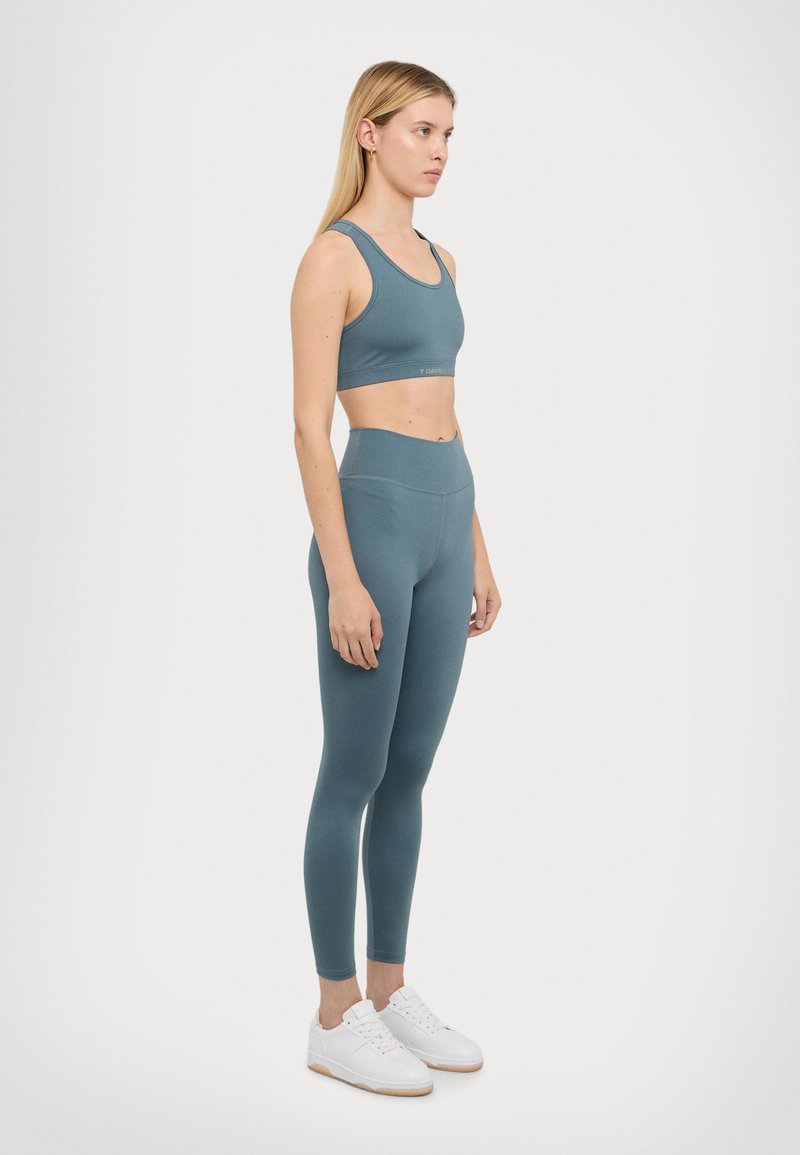 Teal sports bra and matching high-waisted leggings made from stretchy fabric. White sneakers with a textured upper and gum sole.