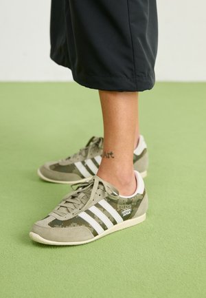 Persoon draagt beige en witte Adidas Japan sneakers op een groene vloer, met een tatoeage met de tekst "Art" op de linker enkel.