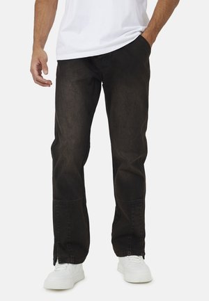 Mann trägt schwarze gerade Hose mit Seitenschlitzen an den Knöcheln, weiße Turnschuhe und ein weißes T-Shirt, Hand in der Tasche.