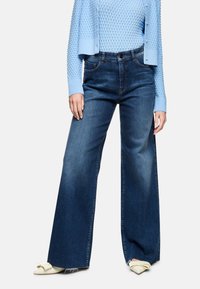Wide-leg dark blue denim jeans with subtle fading, paired with a light blue textured sweater and cream-colored pointed flats.
