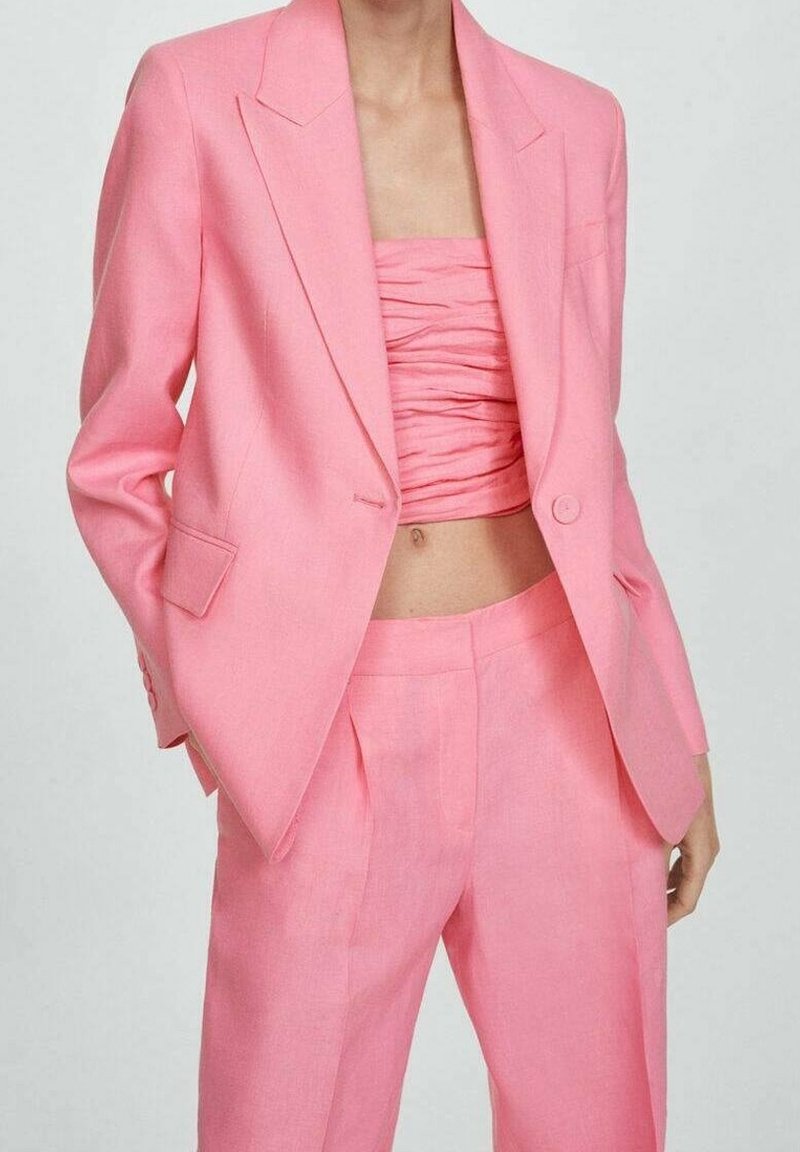 Femme portant un blazer cintré rose vif et un pantalon taille haute assorti, accompagné d'un top bandeau froncé, debout les mains dans les poches.