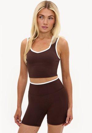 Kvinde med langt blond hår iført matchende mørkebrunt atletisk cropped top og højtaljede cykelshorts med hvide detaljer, stående mod ensfarvet baggrund.