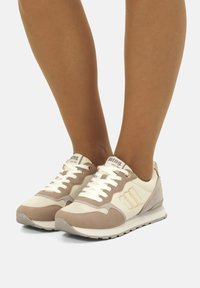 Baskets de sport beiges et crème avec des accents en suède et en mesh, design à lacets, semelle en caoutchouc et détail du logo sur le côté.
