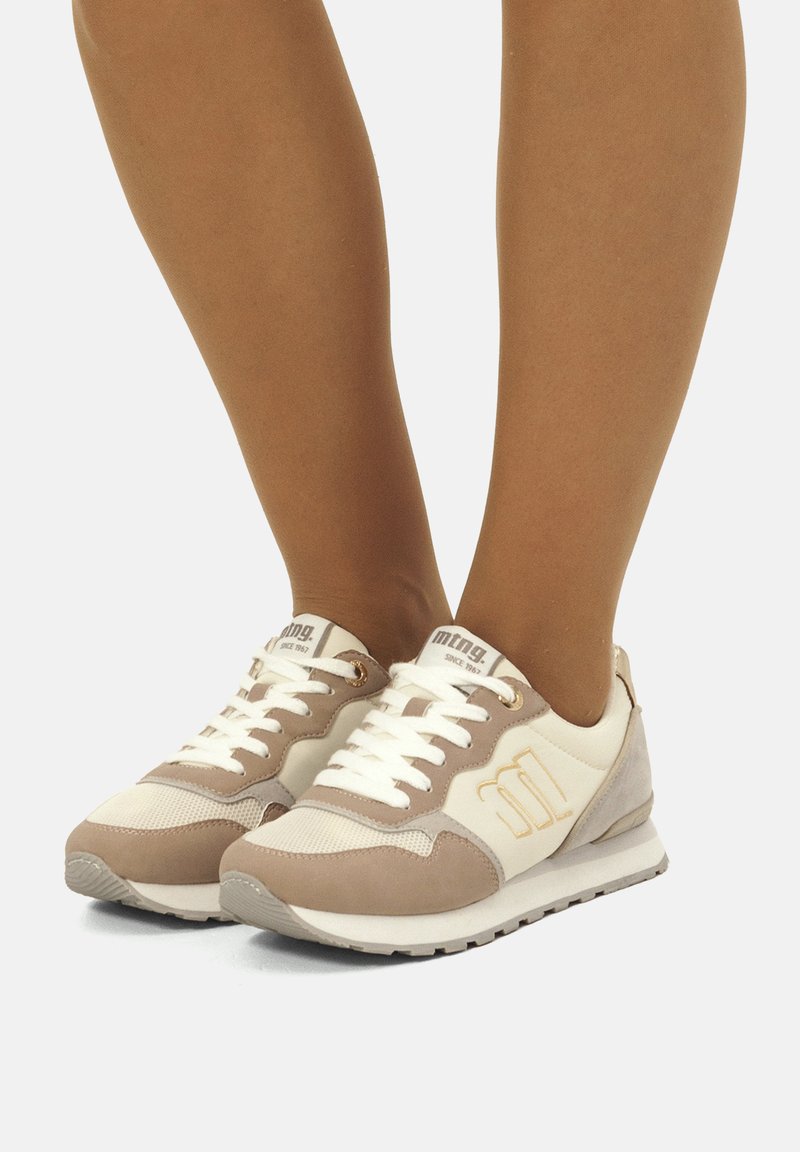 Baskets de sport beiges et crème avec des accents en suède et en mesh, design à lacets, semelle en caoutchouc et détail du logo sur le côté.