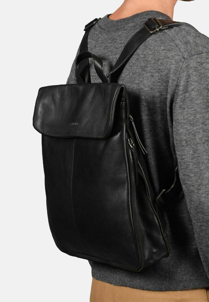 Mochila de cuero negra con tapa plegable, cuenta con cremalleras en los laterales y correas ajustables para los hombros, textura suave y un branding mínimo.