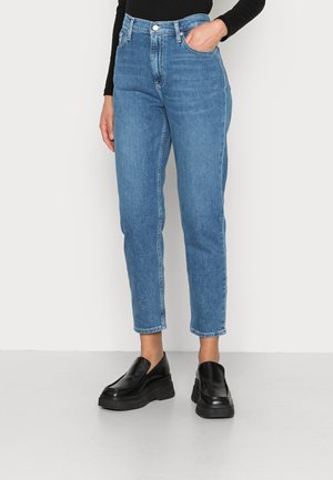 Osoba nosząca niebieskie jeansy z wysokim stanem i czarne slip-on na platformie, z jedną ręką w kieszeni, stojąca na szarej podłodze przy białej ścianie.