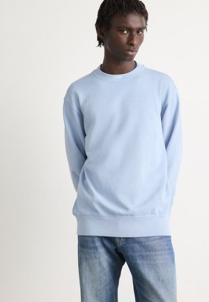 Sudadera de cuello redondo azul claro hecha de algodón suave, con puños y dobladillo de canalé, y con un logo sutil en el cuello. Combinada con unos vaqueros.