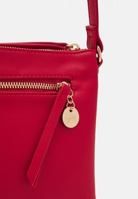 Borsa a tracolla rossa in finta pelle con cerniera dorata, un laccetto in pelle e un'etichetta rotonda dorata con un logo. Texture liscia, design compatto.