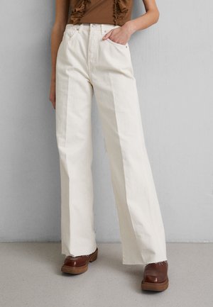 Wide-leg, high-waisted white denim pants with prominent stitched seams and frayed hems, paired with brown wood-soled shoes.