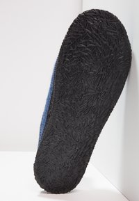 Wollen slipper met een blauwe bovenkant en een getextureerde zwarte rubbersole met een bladpatroon voor grip. Zijaanzicht dat de detail van de zool toont.