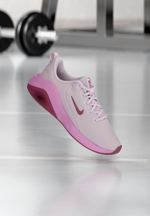 Svetlofialová a ružová športová obuv Nike so šnúrkami plávajúca nad podlahou posilňovne, činky rozmazané v pozadí.