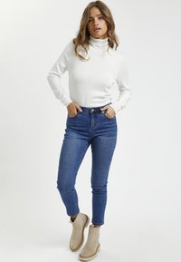 Pull à col roulé blanc, jeans taille haute bleus et bottines beige à semelle en caoutchouc. Texture lisse, design ajusté.