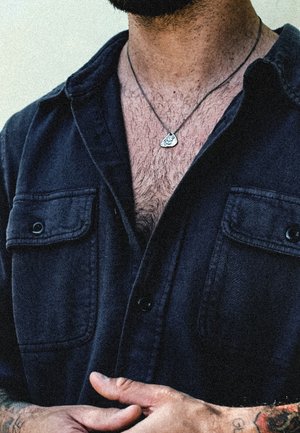 Black button-up shirt with two front pockets, short sleeves, and a silver pendant necklace. Textured fabric and casual fit.