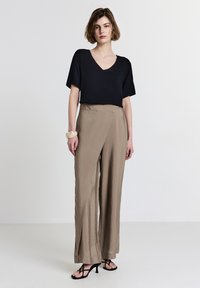 Donna che indossa una maglietta nera a V con maniche corte, pantaloni beige a gamba larga, bracciale beige e sandali neri con tacco, in piedi su uno sfondo bianco.