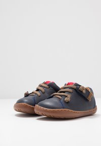 Scarpe in pelle blu scuro con suole in gomma marrone, caratterizzate da lacci marroni, una fascia in Velcro e un'acca rossa sulla linguetta.