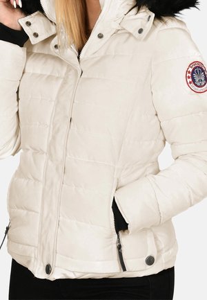 Kobieta w beżowej pikowanej zimowej kurtce z czarnym futrzanym kapturem, zamkiem błyskawicznym i zatrzaskami oraz okrągłą haftowaną łatą na rękawie.