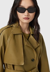 Cappotto trench in pelle verde oliva con un ampio colletto, bottoni neri doppi e una cintura; indossato con occhiali da sole neri a forma rettangolare.