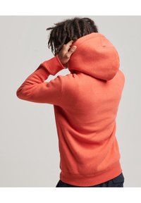 Sudadera naranja con capucha, puños acanalados y una tela texturizada. La persona está ajustando la capucha mientras está de espaldas.