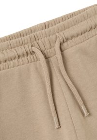 I pantaloni della tuta beige in cotone presentano un elastico a coste in vita e un cordino piatto. La superficie ha una texture liscia con dettagli di cucitura sottili.
