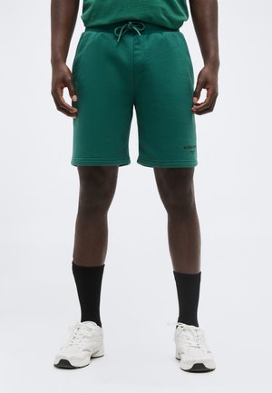 Κάτω μέρος σώματος ενός ατόμου που φοράει πράσινα σορτς με κορδόνι, μαύρες κάλτσες και λευκά αθλητικά παπούτσια, στέκεται μπροστά σε ένα απλό φόντο.