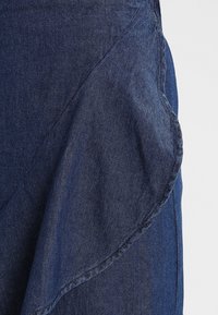 Tessuto denim di colore blu navy scuro con dettaglio a bordo arricciato. Il tessuto ha una texture liscia con cuciture visibili lungo le cuciture.