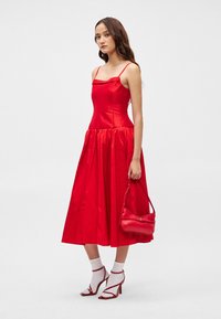 Κόκκινο σατέν φόρεμα με εφαρμοστό κορμό και φουσκωτό φούστα. Συνδυάζεται με κόκκινη τσάντα, λευκές κάλτσες και κόκκινες ψηλοτάκουνες σαγιονάρες.