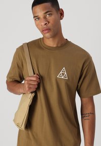 Camiseta de algodón marrón con un gráfico triangular blanco que incluye letras. El modelo sostiene un bolso de hombro beige, mostrando un ajuste relajado.