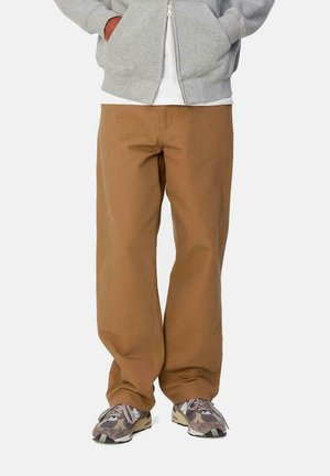 Pantalones anchos marrones de tela de algodón, con una textura suave; llevados con zapatillas grises que presentan un patrón de camuflaje.