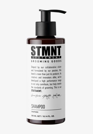 Bouteille brune avec un bouchon-pompe noir. L'étiquette blanche présente un texte noir indiquant "STMNT GROOMING GOODS" et "SHAMPOOING", avec les détails du volume en dessous.