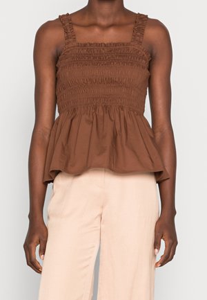 Nainen, jolla on ruskea, röyhelöillä koristeltu, hihaton toppi ja beigehousut, seisoo yksinkertaista vaaleaa taustaa vasten.