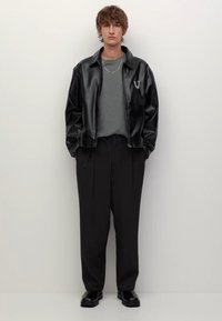 Zwarte leren jas, grijs shirt, losse zwarte broek, glanzende zwarte schoenen. Belangrijke details: kettingaccent op de jas, ontspannen pasvorm.