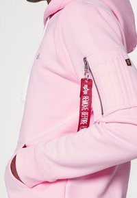 Person som bär en ljusrosa hoodie med en ficka med dragkedja på ärmen och en röd tygnyckelring märkt "REMOVE BEFORE FLIGHT."